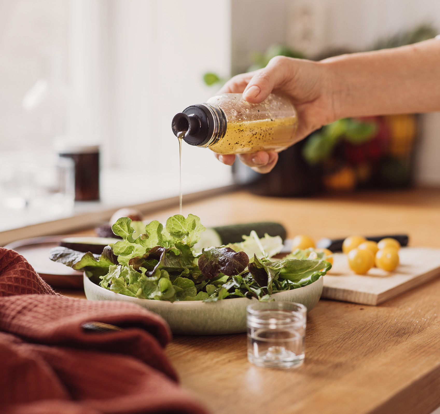 A hand pouring salad dressing onto a salad which sits on a white plate on a table next to a glass and cutting board